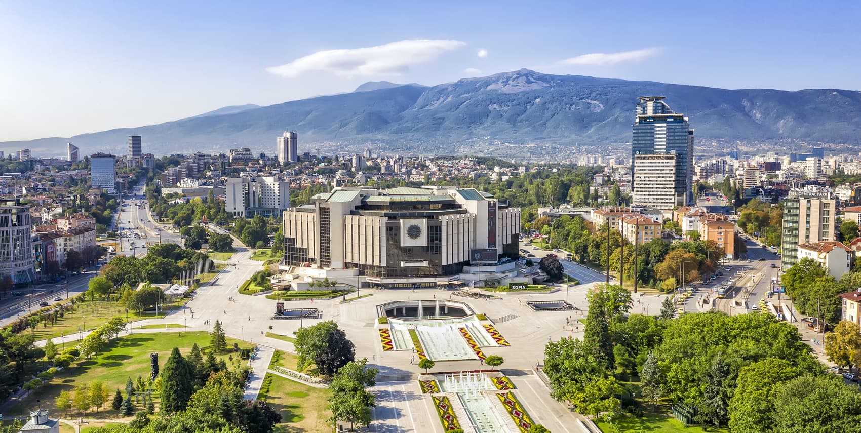Panoramic view of the National Palace of Culture in Sofia, Bulgaria, surrounded by a large park, fountains, and the Vitosha Mountain range in the background.